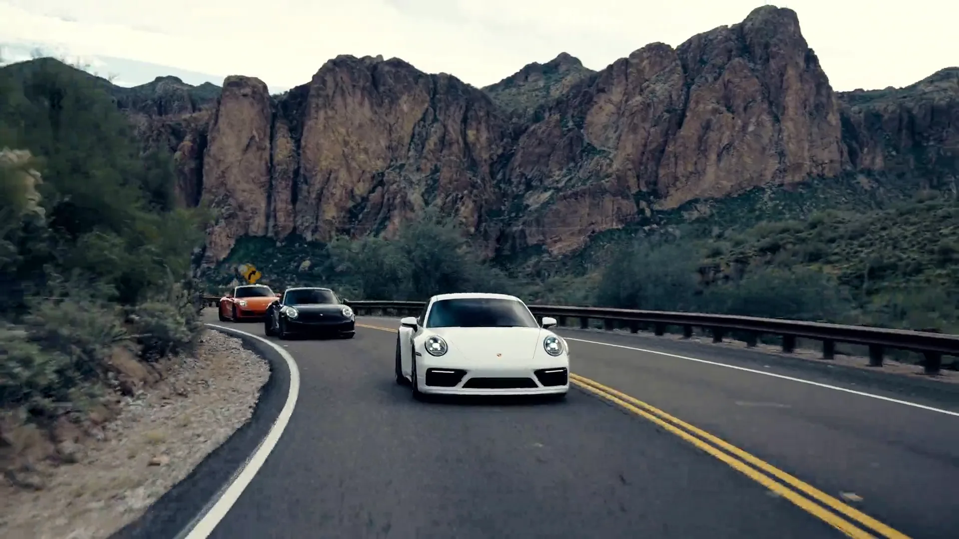 An orange Porsche 911 leading a line of Deluxe Car Club cars through a sweeping bend on an Arizona desert road.